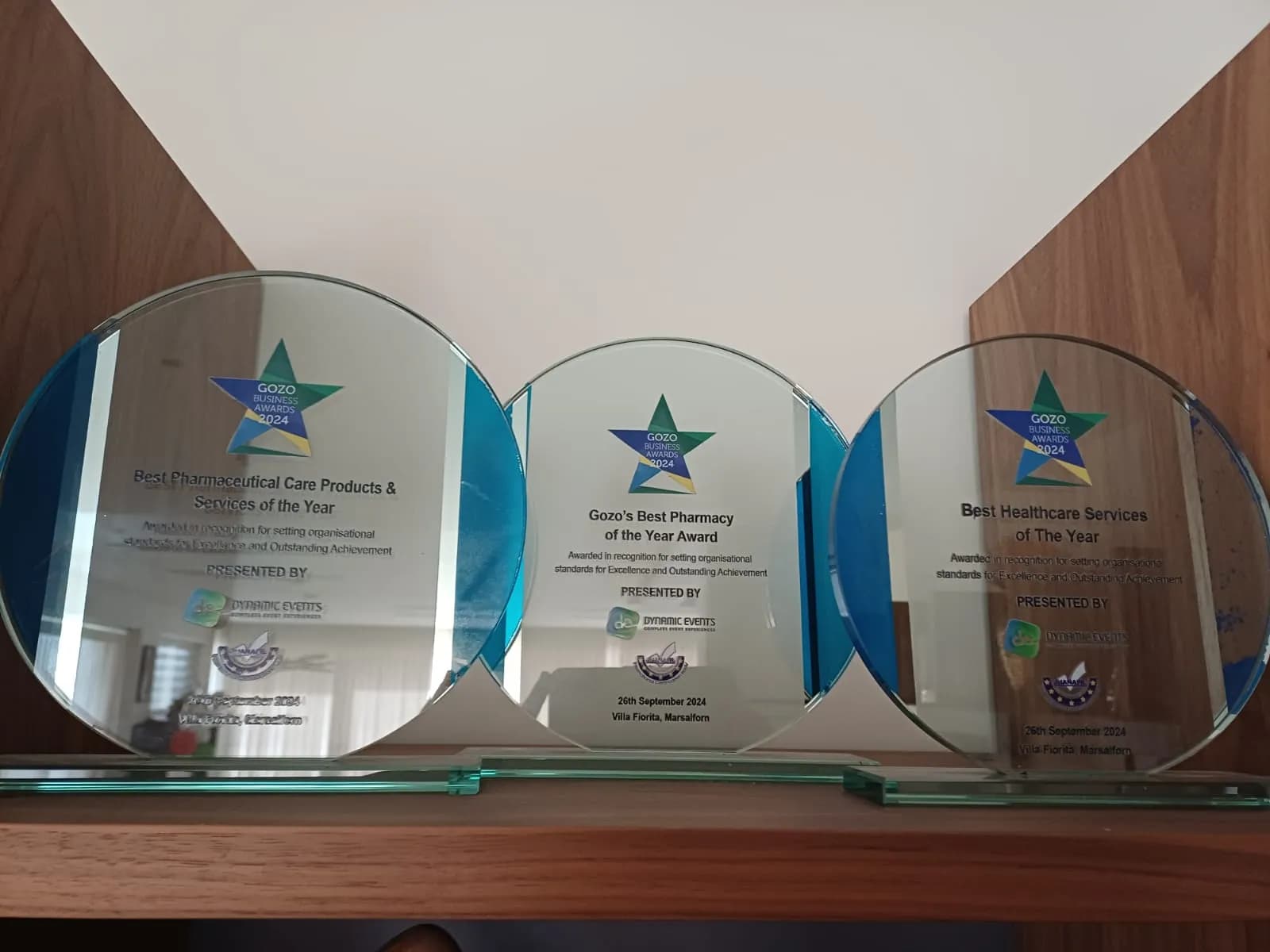 Three Gozo Business Awards 2024 glass trophies on a shelf: Best Pharmaceutical Care Products & Services, Gozo's Best Pharmacy of the Year, and Best Healthcare Services of the Year.